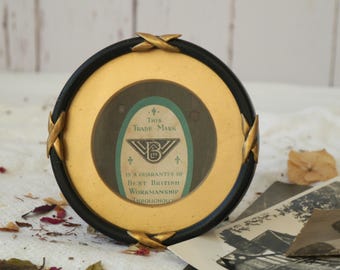 Könnte beinhalten: Ein kleiner, runder Bilderrahmen mit goldfarbenem Rahmen, schwarzem Außenrand und dekorativen Messingakzenten. Der Rahmen hat eine Glasfront und ein Etikett im Inneren mit der Aufschrift "This Trade Mark is a guarantee of Best British Workmanship Throughout".