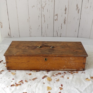 May include: A rectangular wooden storage box with a natural brown finish, featuring a metal handle on the top lid and a central keyhole on the front panel. The box displays visible wood grain patterns and signs of wear along the bottom edges.