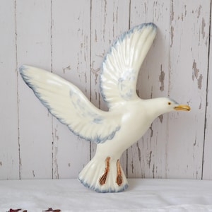 Op de afbeelding: Keramische zeemeeuwsculptuur in vlucht, met uitgestrekte vleugels. De vogel is voornamelijk wit met lichtblauwe accenten op de vleugels en staart. De snavel is geel en de poten bruin. De sculptuur staat tegen een witte, verweerde houten achtergrond.