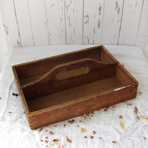 Puede incluir: Una caja organizadora rectangular de madera con acabado marrón oscuro y un divisor central que incluye un asa integrada. La caja presenta signos visibles de desgaste, como pintura desconchada en los bordes, y paredes rectas.
