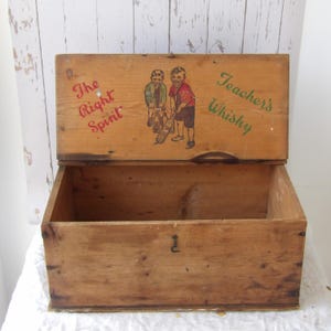 May include: Open wooden box with a hinged lid. The lid features the text "The Right Spirit" and "Teacher's Whisky" with an illustration of two boys. The box is made of light-coloured wood.
