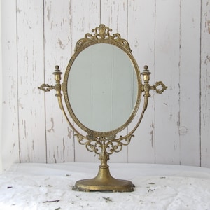 Op de afbeelding: Een antieke messing toilettafelspiegel met een ovaalvormige spiegel en sierlijke details. De spiegel wordt ondersteund door een decoratieve voet en heeft ingewikkeld krulwerk en bloemmotieven. De spiegel is ongeveer 30 cm hoog.