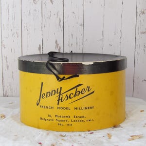 May include: A vintage yellow hat box with a black lid and trim. The box features the text "Jenny Fischer FRENCH MODEL MILLINERY" in black script. The box is cylindrical and has a black ribbon tied around it.