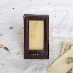 May include: A rectangular picture frame with a dark brown, smooth finish and a bevelled edge design. The frame features a light cream-coloured, textured insert with a subtle herringbone pattern. The frame has a vertical orientation and a simple border.