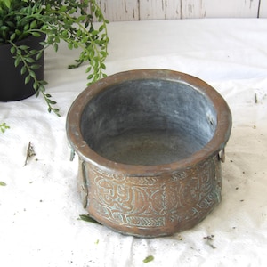 May include: A round, antique copper pot with etched floral designs. The pot has a dark interior and a weathered, aged appearance. The pot is sitting on a white surface, with a green plant in a black pot in the background.