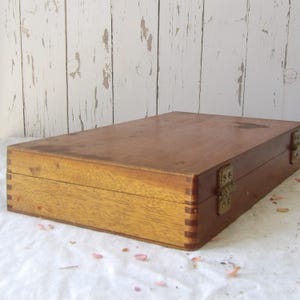 May include: A rectangular wooden box with a hinged lid and metal clasps. The box is made of light and dark brown wood, with visible dovetail joints. The box is sitting on a white surface.