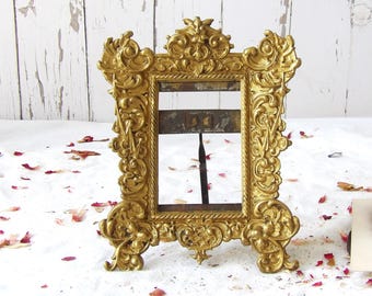 May include: Ornate gold-colored picture frame with intricate scrollwork and a rectangular opening. The frame has a stand for tabletop display. The background is a white surface with scattered rose petals.
