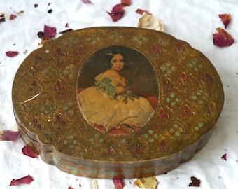 May include: An antique oval-shaped box with a portrait of a woman in a yellow dress. The box has a gold-colored finish with floral and leaf patterns in red and green. The box is on a white surface with scattered rose petals.