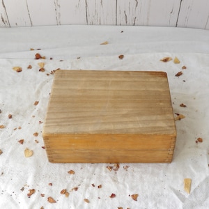May include: A rectangular wooden box with a natural light brown finish and visible wood grain. The box features finger joint construction on the corners and a flat, smooth lid that sits flush with the base, creating a simple, clean appearance.
