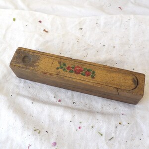 Op de afbeelding: Een rechthoekige houten kist met een schuifdeksel. De kist is geel geverfd en versierd met een bloemenontwerp van rode rozen en groene bladeren. De kist lijkt vintage of antiek te zijn, met tekenen van slijtage.