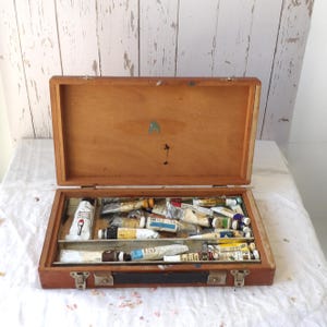 May include: Open wooden artist's paint box with a hinged lid. The box is filled with various tubes of paint in different colors. The box is made of light brown wood and has metal clasps.