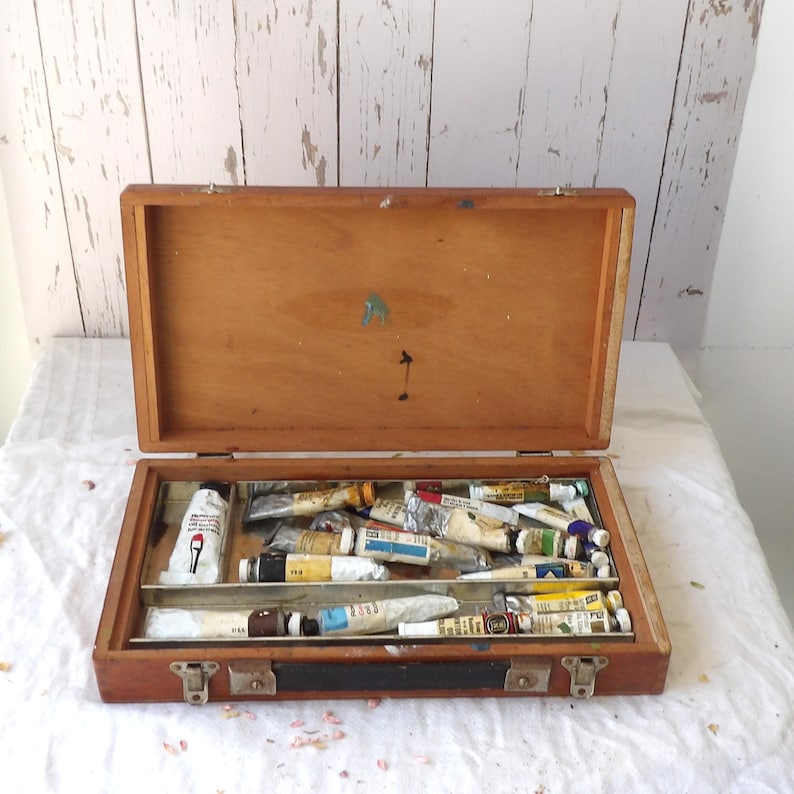 May include: Open wooden artist's paint box with a hinged lid. The box is filled with various tubes of paint in different colors. The box is made of light brown wood and has metal clasps.