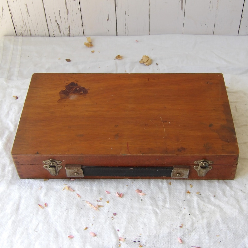 May include: A rectangular wooden box with a dark brown finish. The box has metal clasps and a black leather handle. The wood shows signs of wear and tear, with some stains on the surface. The box is closed and appears to be vintage.