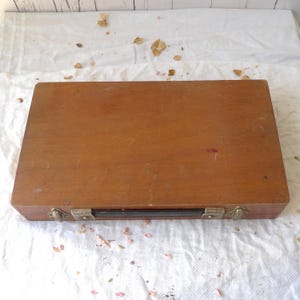 May include: A rectangular wooden box with a dark brown finish. The box has metal clasps and a black handle. The box is closed and sits on a white cloth surface. The wood grain is visible on the box.