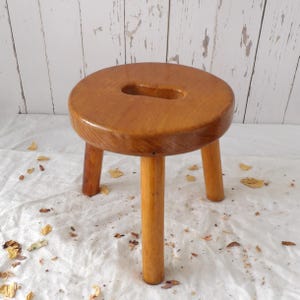 Peut inclure: Un petit tabouret rond en bois brun clair avec trois pieds cylindriques et une finition lisse et polie. L'assise circulaire présente une poignée centrale découpée en forme d'ovale allongé, offrant une prise fonctionnelle pour le transport.