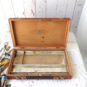 May include: An open, vintage wooden artist's paint box with a metal tray. The box is made of light brown wood and has metal clasps and a handle. Paint tubes are visible in the lower left corner.
