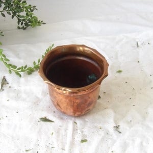 Puede incluir: Una pequeña maceta de cobre antigua con un borde festoneado. El interior es oscuro y el exterior tiene una cálida pátina marrón rojiza. La maceta está vacía y se encuentra sobre una superficie blanca, con follaje verde en el fondo.