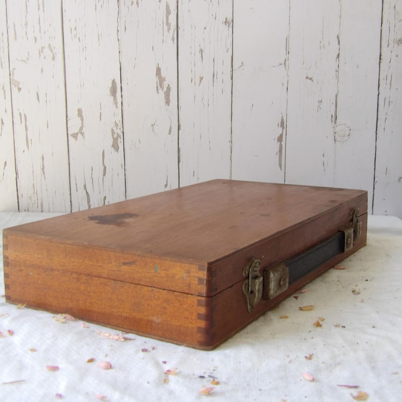 May include: A rectangular wooden box with a dark brown finish. The box has metal clasps and a black leather handle. The box is sitting on a white surface in front of a white wooden background.