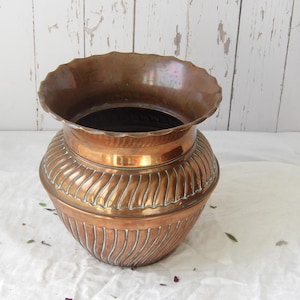 Puede incluir: Una jardinera de cobre vintage con un borde ancho y festoneado y un cuerpo bulboso. La jardinera presenta detalles verticales y acanalados alrededor de la base y un acabado pulido. El interior es oscuro, contrastando con el exterior de cobre brillante. Una pieza decorativa.