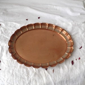 May include: An oval copper-coloured tray with a scalloped edge. The tray has a smooth, reflective surface and a decorative border. The tray is empty and sits on a white surface. The tray is likely used for serving or display.