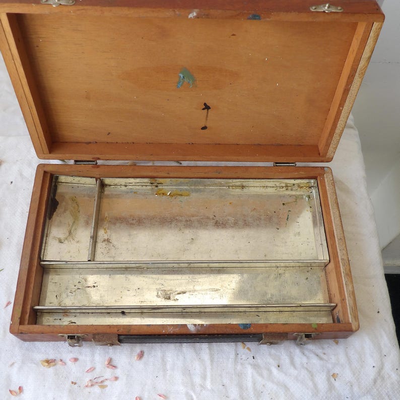 May include: An open wooden artist's paint box with a hinged lid. The interior features a metal palette with several compartments for mixing paints. The box is made of light-colored wood and shows signs of use, with paint splatters.