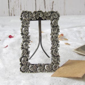 Peut inclure: Un petit cadre photo rectangulaire argenté avec des détails floraux tourbillonnants ornés. Le cadre a un simple support en métal à l'arrière. Le cadre est vide, prêt à afficher une photographie. Le cadre est probablement vintage ou antique.