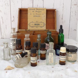Könnte beinhalten: Eine Sammlung antiker Apothekerflaschen und -gläser, einige mit Etiketten, angeordnet vor einer offenen Holzkiste. Die Flaschen variieren in Größe, Form und Farbe, mit dunkelbraunem und klarem Glas. Ein Vintage-Schild ist sichtbar.