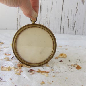May include: A circular picture frame with a metallic gold-toned finish and a smooth, rounded edge. The frame features a small, integrated loop at the top for hanging or display purposes, surrounding a plain, off-white, circular centre insert.