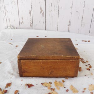 May include: A square wooden box with a natural light brown finish and a flat lid. The box features visible wood grain patterns and a weathered, worn texture on the surface, showing signs of age and use across its exterior panels.
