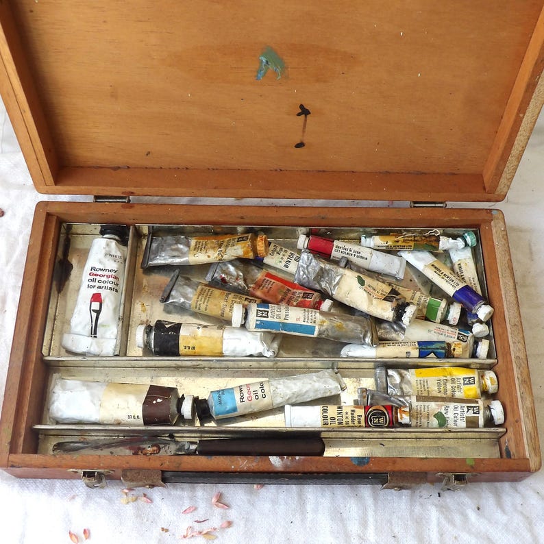 May include: Open wooden artist's paint box filled with various oil paint tubes in a range of colors. The box has metal trays and a palette knife. The lid is open, revealing paint splatters. The text on one tube reads "Rowney Georgian oil colour for artists."