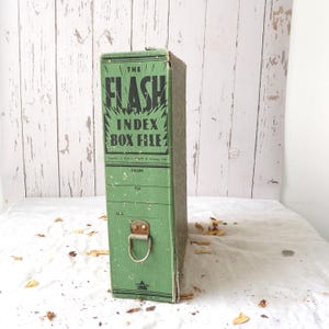 May include: A green cardboard index box file with black text reading THE FLASH INDEX BOX FILE. The spine features a metal pull handle, lines for labelling FROM and TO, and a small triangular logo at the bottom. The surface has a speckled texture.