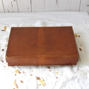 May include: A rectangular, tan-colored leather box with a smooth, flat lid and visible stitching along the edges. The surface of the leather shows natural variations in tone and texture, with a faint, dark stamp visible on the top left corner.