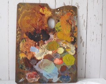 May include: A rectangular wooden artist palette featuring a thumb hole at the top. The surface is covered in thick, dried oil paint in various colors including ochre, brown, black, yellow, green, blue, red, and white, showing heavy texture.
