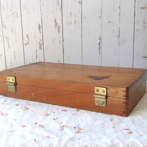 May include: A rectangular wooden box with a warm brown finish. The box features brass-colored hinges and clasps. The wood grain is visible, and there is a dark stain on the top surface. The box is resting on a white surface.