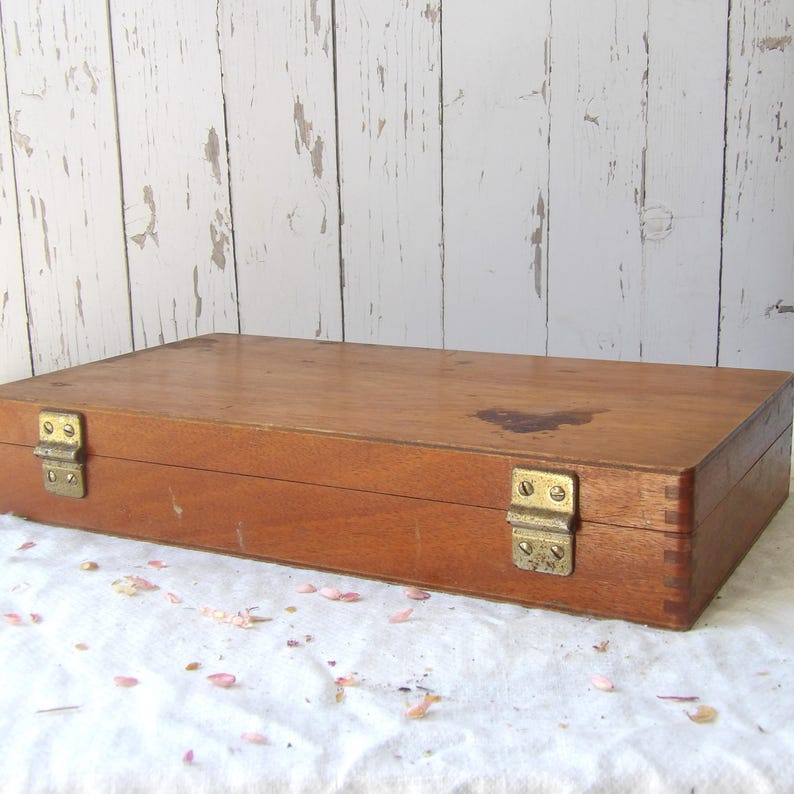 May include: A rectangular wooden box with a warm brown finish. The box features brass-colored hinges and clasps. The wood grain is visible, and there is a dark stain on the top surface. The box is resting on a white surface.
