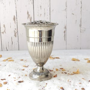Könnte beinhalten: Eine silberfarbene Blumen-Vase aus Metall mit polierter Oberfläche, einem ausgestellten Rand und einem abnehmbaren Gittereinsatz. Der Körper der Vase zeigt vertikale Rillen, während der runde Sockel mit einem blattartigen Rand verziert ist.