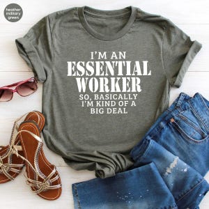 May include: A heather military green short-sleeve t-shirt with white text that reads I'M AN ESSENTIAL WORKER SO, BASICALLY I'M KIND OF A BIG DEAL. The shirt is displayed alongside a pair of blue denim jeans and brown strappy sandals.