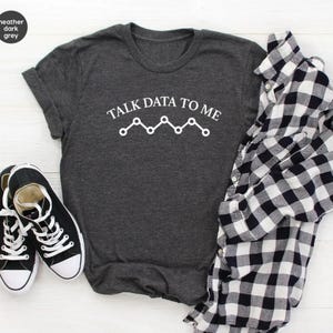 May include: A heather dark grey short-sleeve t-shirt with the white text TALK DATA TO ME and a line graph icon. Beside the shirt is a black and white checkered button-down shirt and a pair of black and white low-top canvas sneakers.
