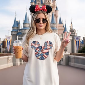 Peut inclure: Un t-shirt couleur crème arborant un grand motif central en forme de silhouette d'oreilles de souris, rempli d'un patchwork d'étoiles et de rayures rouges, blanches et bleues. Il est porté avec un serre-tête à oreilles noires et un nœud rouge à pois.