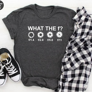 May include: Dark grey t-shirt with the phrase "WHAT THE f?" and camera aperture symbols. The shirt is accompanied by black trainers and a black and white checked shirt.