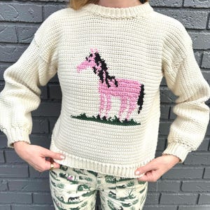 May include: A cream-colored crocheted sweater with a pink horse design. The horse has a black mane and tail.