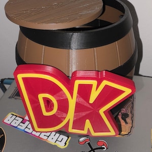 Puede incluir: Un barril marrón con tapa extraíble y logotipo "DK" rojo y amarillo. El barril tiene bandas negras y está sobre un portátil con pegatinas. La tapa es marrón con un diseño de vetas de madera.