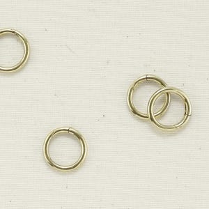 May include: Several small, gold-colored metal rings are scattered on a light-colored fabric surface. The rings are circular with a hinged clasp, suggesting they are designed to open and close. The overall aesthetic is simple and minimalist.