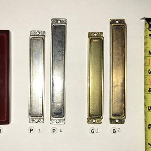 May include: A collection of rectangular hardware pulls in various finishes, including dark brown, silver, and brass. The pulls vary in length, with the longest measuring approximately 6 inches. Each pull has a recessed center and mounting holes.