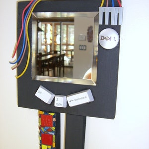 May include: A black framed mirror with a silver trim, featuring a reflection of a room with a window and furniture. The mirror is attached to a black stand with a mosaic tile design on the front. The mirror has computer keys attached to the bottom, including Ctrl, 3, PgDn, and Backspace. The mirror also has a gray electrical outlet and a white round sticker with the word "DELL" on it.