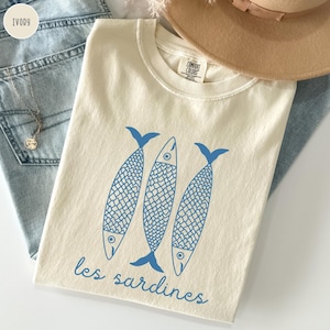 Op de afbeelding: Een cr&egrave;mekleurig T-shirt met een blauwe afbeelding van drie vissen en de tekst "les sardines".