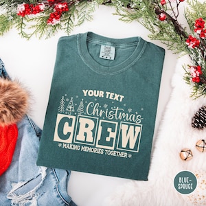以下が含まれることがあります： 「YOUR TEXT Christmas CREW MAKING MEMORIES TOGETHER」の白のレトロスタイルフォントの、ティール色のTシャツ。クルーネックで「Comfort Colors」のタグが付いています。クリスマス装飾がTシャツを囲んでいます。