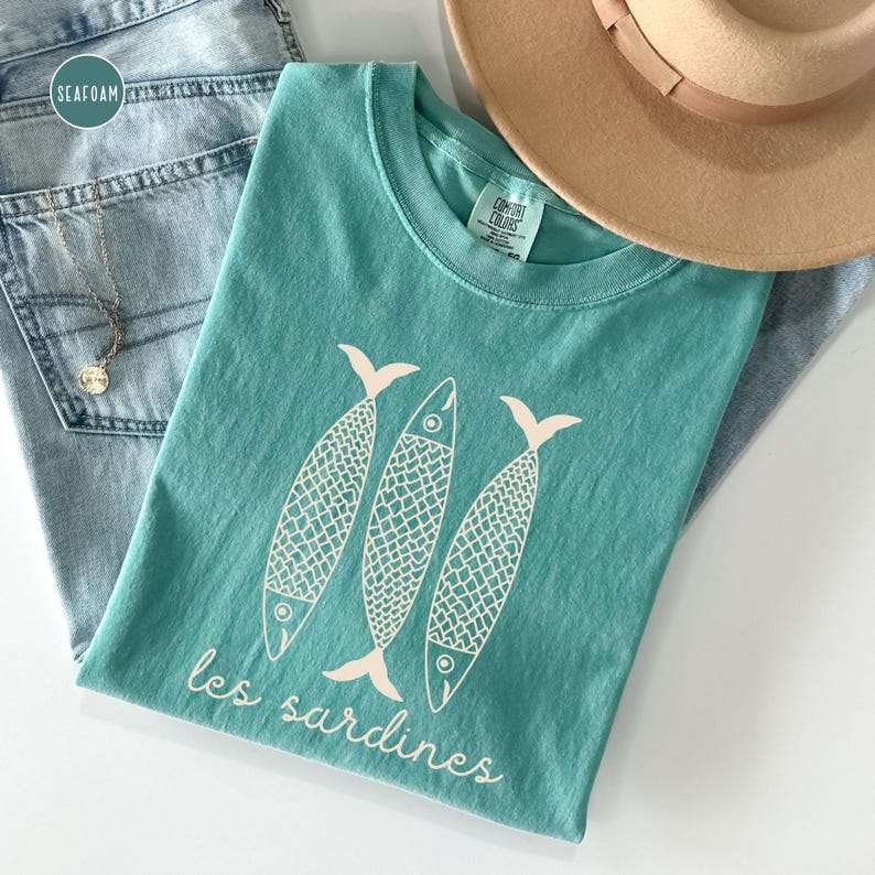 Op de afbeelding: Een turquoise t-shirt met een witte afbeelding van drie gestileerde vissen en de tekst "les sardines".
