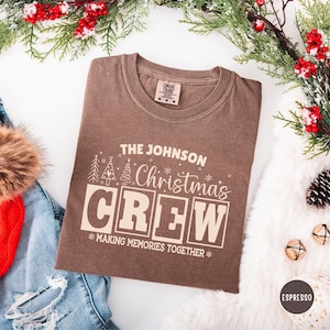 以下が含まれることがあります： お祝いのデザインで「THE JOHNSON Christmas CREW」と書かれた茶色のTシャツ。シャツには「MAKING MEMORIES TOGETHER」というフレーズが含まれており、クリスマスの装飾に囲まれています。
