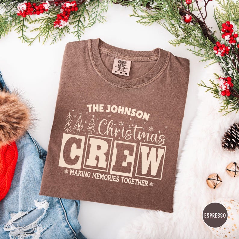 以下が含まれることがあります： お祝いのデザインで「THE JOHNSON Christmas CREW」と書かれた茶色のTシャツ。シャツには「MAKING MEMORIES TOGETHER」というフレーズが含まれており、クリスマスの装飾に囲まれています。